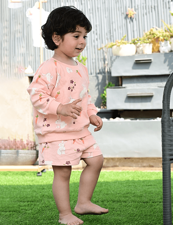 A young child, who appears to be of South Asian descent, walking barefoot wearing a pink organic cotton sweatshirt and shorts with a bunny and carrot print. Shop cute baby clothes India.
