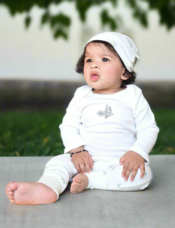 Adorable baby, who appears to be of South Asian descent, sitting outdoors wearing a soft 100% organic cotton bodysuit, leggings, and cap set. Gentle for new skin. Shop India.