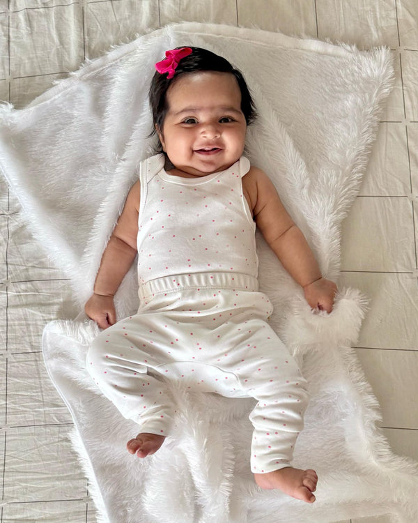 A happy baby, who appears to be of South Asian descent, lying on a soft blanket wearing a white sleeveless bodysuit with small dots. Shop comfortable baby vests India.