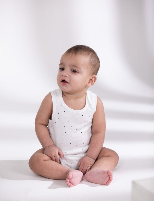 A sweet baby, who appears to be of South Asian descent, sitting on a white surface wearing a white sleeveless bodysuit with very small dots. Shop comfortable baby vests India.