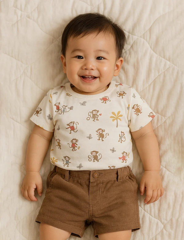 Adorable baby, who appears to be of East Asian descent, wearing a 100% organic cotton monkey print t-shirt and warm brown shorts. Shop organic baby sets India.