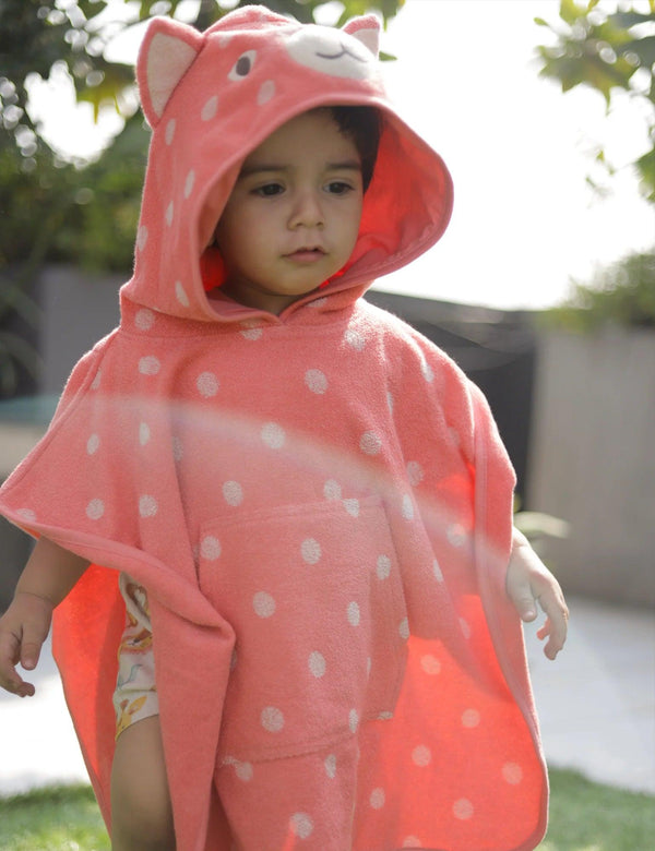 A young child, who appears to be of Caucasian descent, wearing a coral pink organic cotton hooded towel with white polka dots and cute cat ear details. Shop gentle baby towels India.