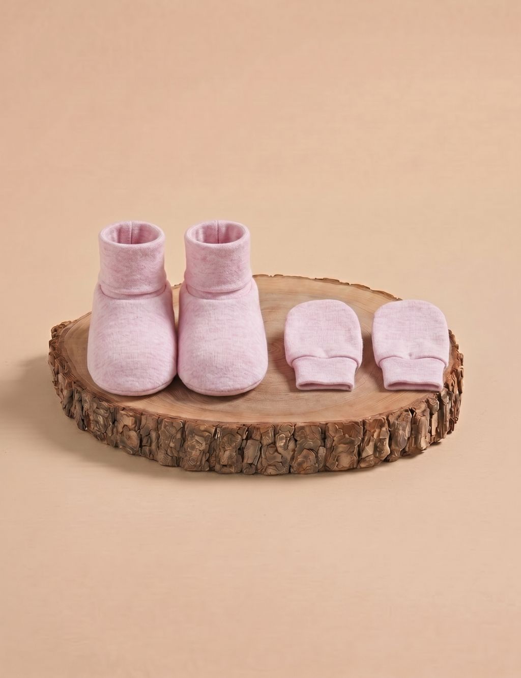 A matching set of solid light pink baby booties and scratch mittens, displayed on a rustic wooden log slice against a plain beige background.