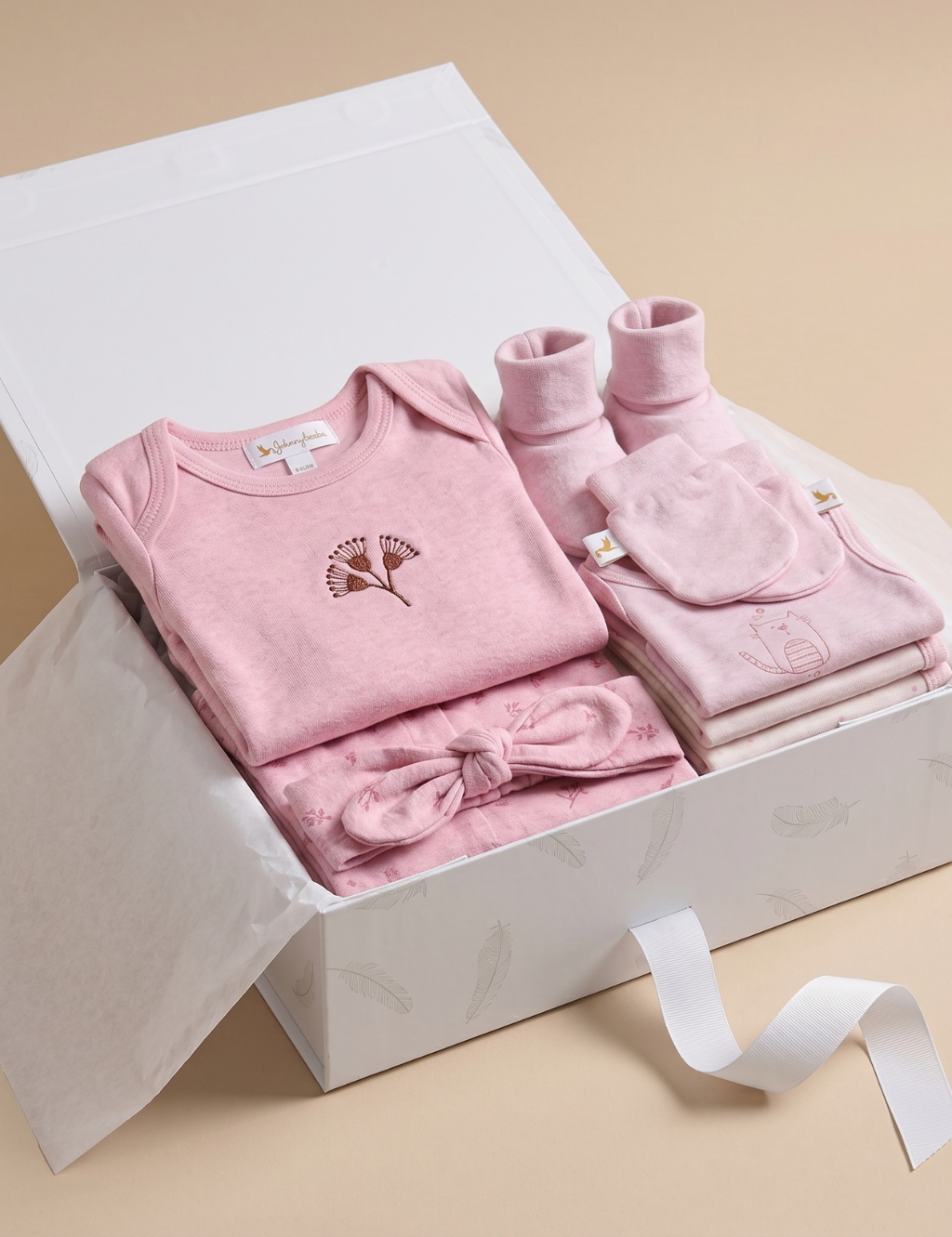 An open white gift box lined with tissue paper, presenting a neatly arranged collection of light pink baby clothing and accessories. The set features a pink long-sleeve bodysuit with a small floral embroidery on the chest, a matching knotted headband, solid pink booties and scratch mittens, and a stack of folded pink garments, all shown against a plain beige background.