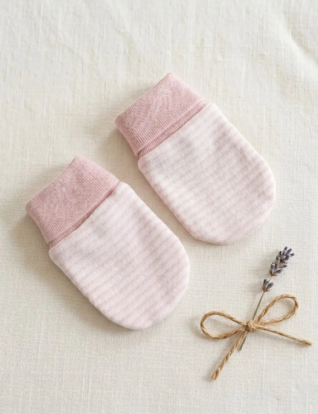 A pair of light pink baby scratch mittens featuring thin horizontal stripes and solid pink ribbed cuffs, laid flat on a textured cream fabric surface alongside a small sprig of lavender tied with twine.