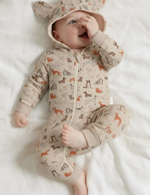 Smiling baby in beige quilted romper with hood featuring 3D ears and dog print pattern, lying on white bed