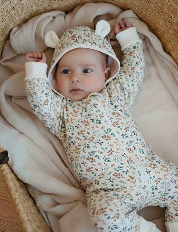 Baby wearing a floral hooded romper with fabric ears, lying in a woven basket with beige muslin blankets