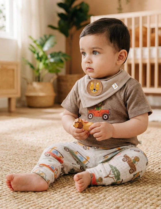 Baby boy model sitting on a rug wearing a safari car t-shirt, printed leggings, and lion patch bandana bib.