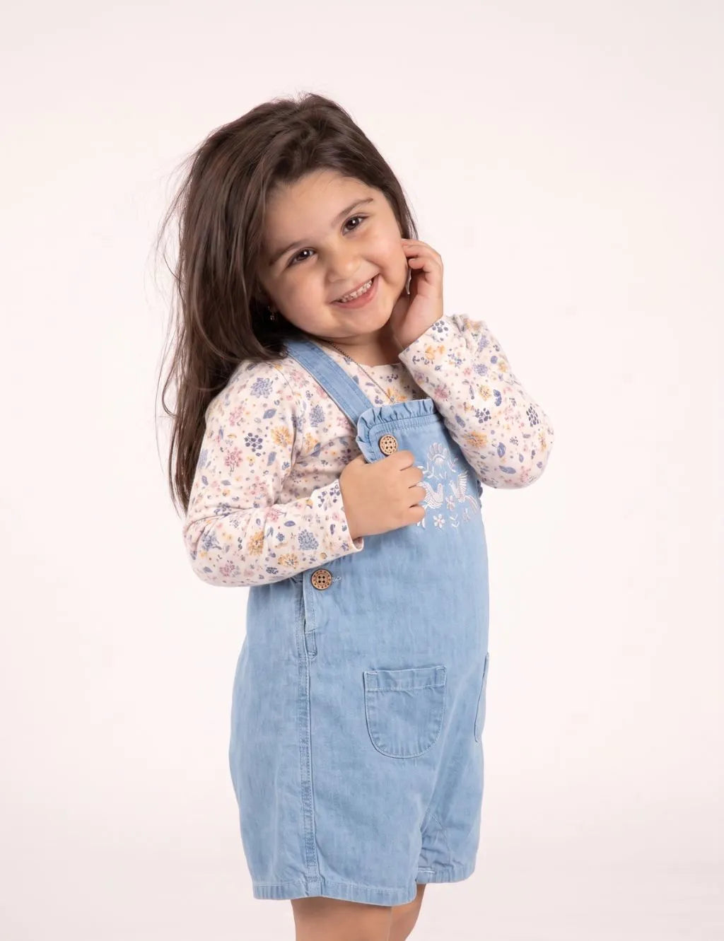 Side view of a smiling toddler girl posing in a light wash denim embroidered short romper over a floral top.