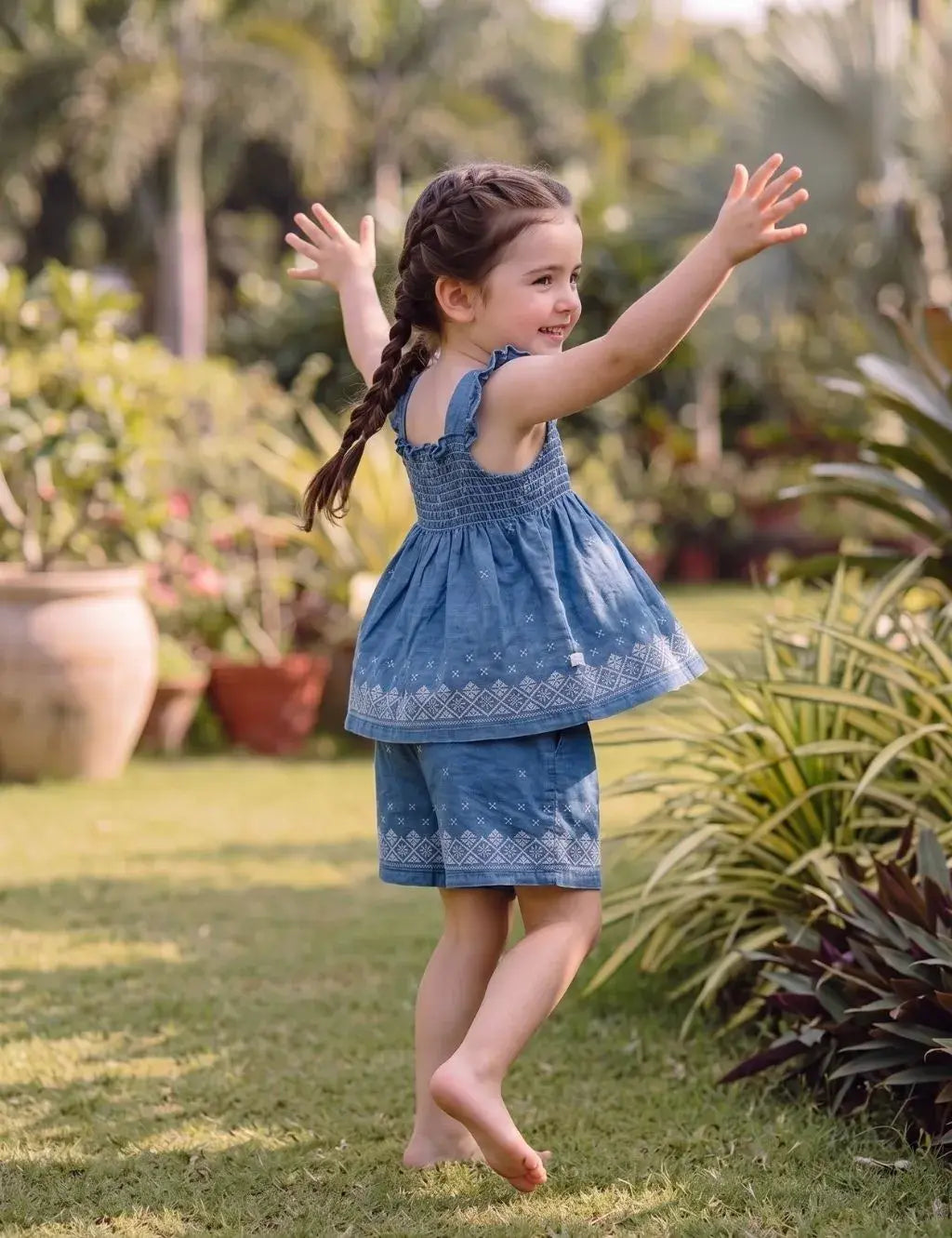 Young girl model outdoors in a garden with arms out, wearing an organic cotton denim blue smocked top and shorts set with white embroidery.