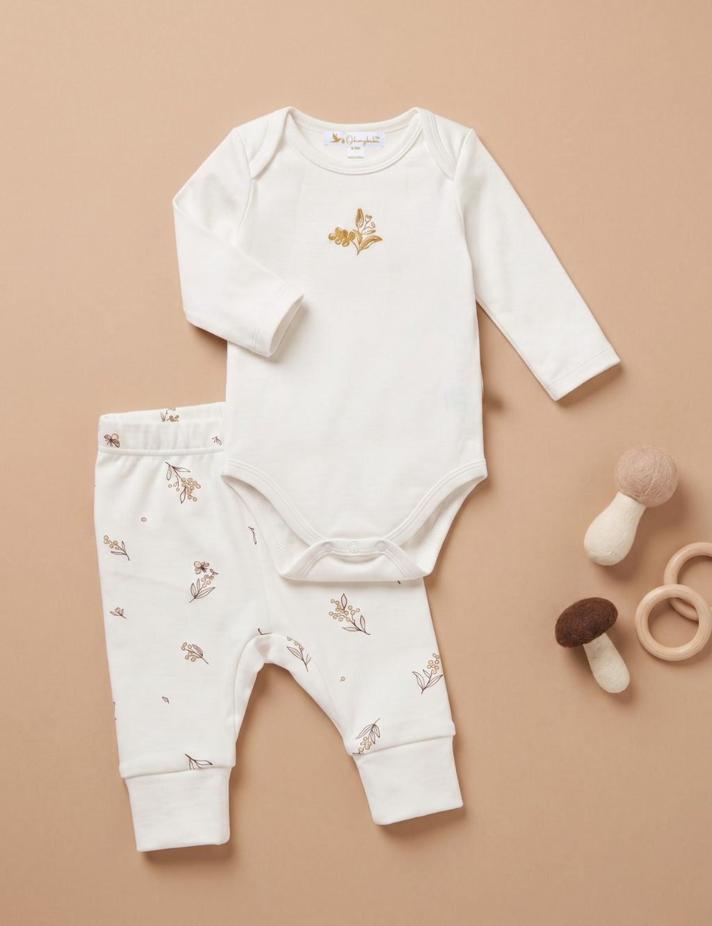 Flat lay view of a baby clothing set featuring a long-sleeve white bodysuit with a small gold floral embroidery on the chest, paired with white pants patterned with delicate gold flowers. The clothes are displayed next to soft mushroom toys and wooden rings against a plain beige background.