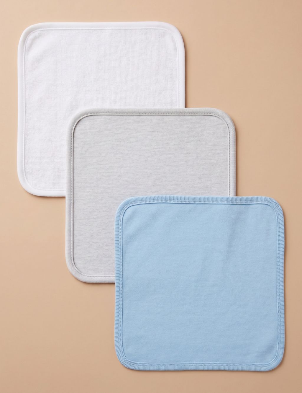 Flat lay view of three square baby washcloths arranged diagonally. From top to bottom: a solid white terry cloth washcloth, a solid light grey heather washcloth, and a solid light blue washcloth, all with soft rounded corners and neat stitched borders, displayed against a plain beige background.